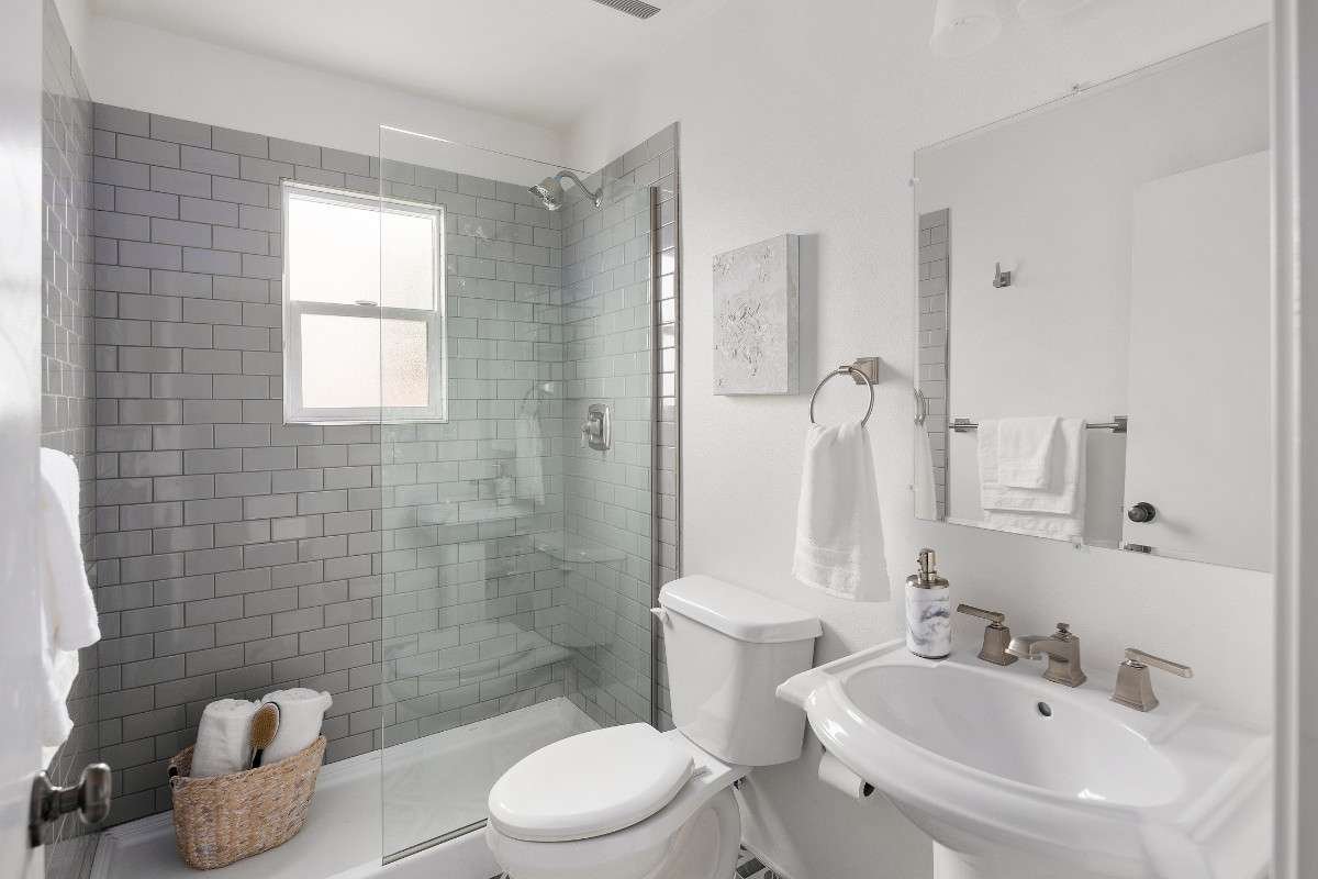 A modern white bathroom featuring a glass-enclosed shower with grey subway tiles, a pedestal sink, and a toilet, all in a clean, minimalist design.