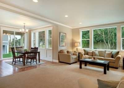 An open-plan living and dining area with beige carpeting, a dining table set, and a kitchen visible in the background.