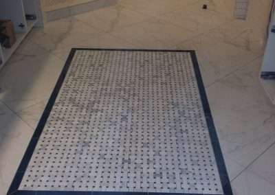 A rectangular floor mosaic made of small black and white tiles, bordered by a darker frame, set within large marble tiles in a bathroom.