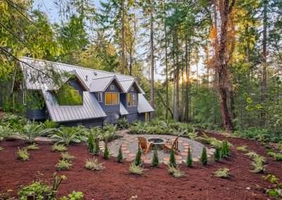 A landscaped backyard with a fire pit area and seating, surrounded by tall trees and lush greenery.