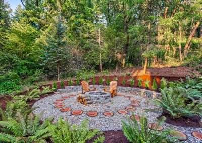 A modern cabin with a landscaped backyard and fire pit area, surrounded by tall trees and lit windows at dusk.