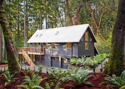 A modern cabin in the forest with a white metal roof, wooden deck, and lush greenery surrounding it.