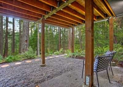 A covered patio area under the cabin with a chair, surrounded by forest vegetation and tall trees.