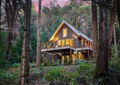 The side view of a modern cabin with a metal roof and a wooden deck, surrounded by tall trees and forest vegetation.