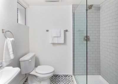 A compact modern bathroom featuring a glass-enclosed shower with grey subway tiles, a white toilet, and patterned tile flooring.