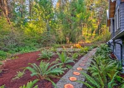A landscaped backyard with a gravel pathway lined with wooden stepping stones, leading to a fire pit area surrounded by tall trees and ferns.
