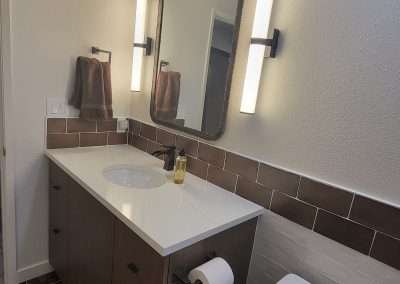 Full view of a bathroom showing vanity, mirror, vertical light fixtures, toilet, and brown accent tiles.