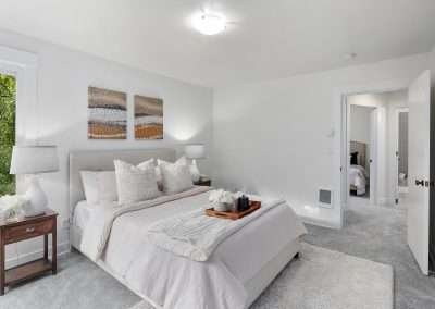 A bright guest bedroom with a plush bed, bedside lamp, and windows overlooking the forest, featuring soft carpeting.