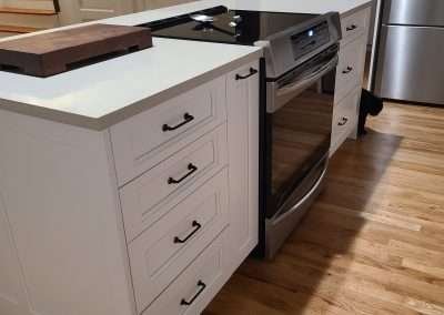 A contemporary kitchen island with a cooktop, white drawers with black handles, and a cutting board on top.