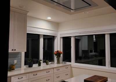 A modern kitchen island with an integrated cooktop and overhead vent hood, surrounded by white cabinetry and large windows.