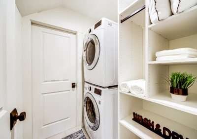 A bright laundry room with stacked washer and dryer units, shelving, and storage space for linens.