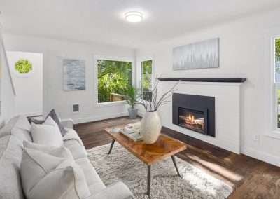 A living room featuring a modern fireplace, light grey couch, wooden coffee table, and large windows overlooking greenery