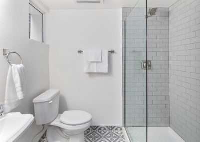 A modern bathroom with a bathtub, shower, toilet, and pedestal sink, featuring white tiles and a wooden shelf.