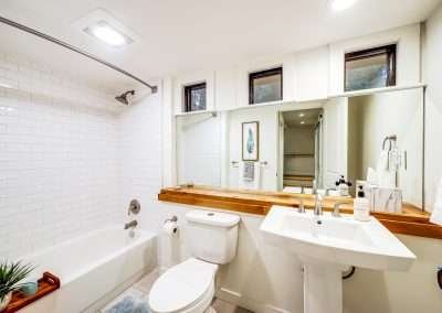 A modern bathroom with a bathtub, shower, toilet, and pedestal sink, featuring white tiles and a wooden shelf.
