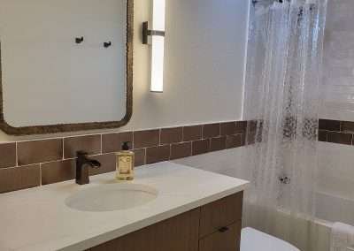 A modern bathroom featuring a white vanity with brown cabinets, a large mirror, vertical light fixture, and clear shower curtain.
