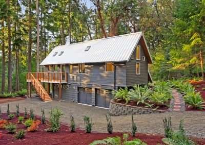 The front exterior of a modern cabin with a white metal roof, wooden deck, and gravel driveway, set in a forest.