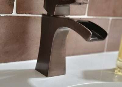 Close-up of a modern dark bronze bathroom faucet with a waterfall spout, set against brown tiled backsplash and white sink.