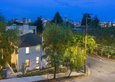 A view of a cityscape at dusk, with a residential neighborhood in the foreground, surrounded by trees and illuminated buildings in the background.