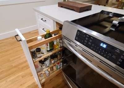 A convenient pull-out spice rack located next to the stove in a modern kitchen with white cabinets and black handles.