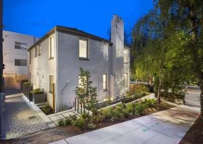 A rear view of the renovated home with modern fencing, a paved walkway, and adjacent structures.