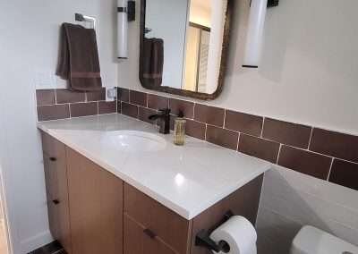 Close-up of a vertical light fixture, brown tile backsplash, and part of a decorative mirror frame in a bathroom.