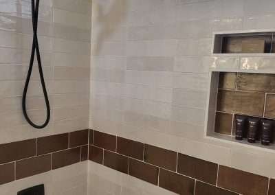 Detail of a shower area showing a black shower head, white and brown tiled walls, and built-in shelving.