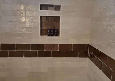 Close-up of a shower area with white tile, brown accent strip, built-in shelving niche, and glass block window.