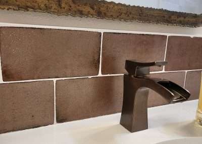 A close-up view of a modern bathroom faucet with a rustic finish, set against brown textured tiles and a white countertop.