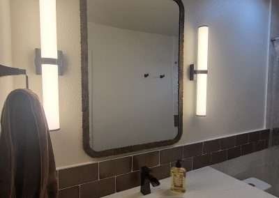 A modern bathroom vanity featuring a sleek countertop, a round sink, a decorative mirror with two vertical light fixtures, and brown accent tiles.