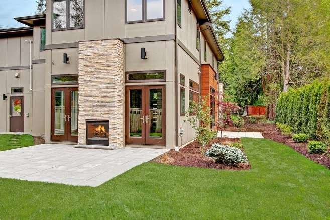 A modern home exterior featuring a stone outdoor fireplace, large glass doors, and a well-manicured lawn surrounded by greenery.