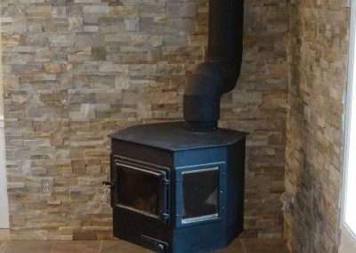 A black wood stove in a corner with a stone wall and tiled floor.