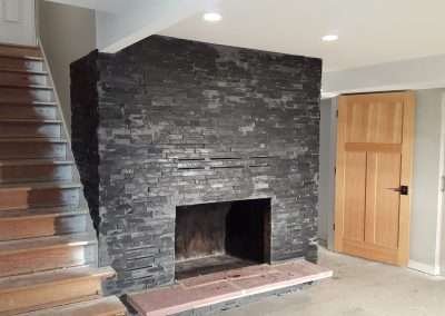 Black stone fireplace in a modern basement with white walls and wooden stairs.