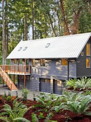 A modern cabin in the forest with a white metal roof, wooden deck, and lush greenery surrounding it.