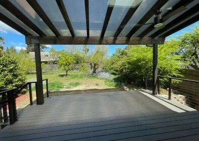 Modern covered patio with a transparent pergola roof, black metal railing, and composite decking, offering a scenic backyard view in Seattle. Designed for style and durability by expert patio installers in Seattle.