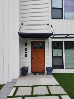 Modern home entrance with a wooden door, black canopy, white siding, and a landscaped walkway featuring geometric grass and stone pavers.