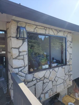 Exterior view of a house with a large window framed by rustic stone cladding, reflecting greenery from the outside.