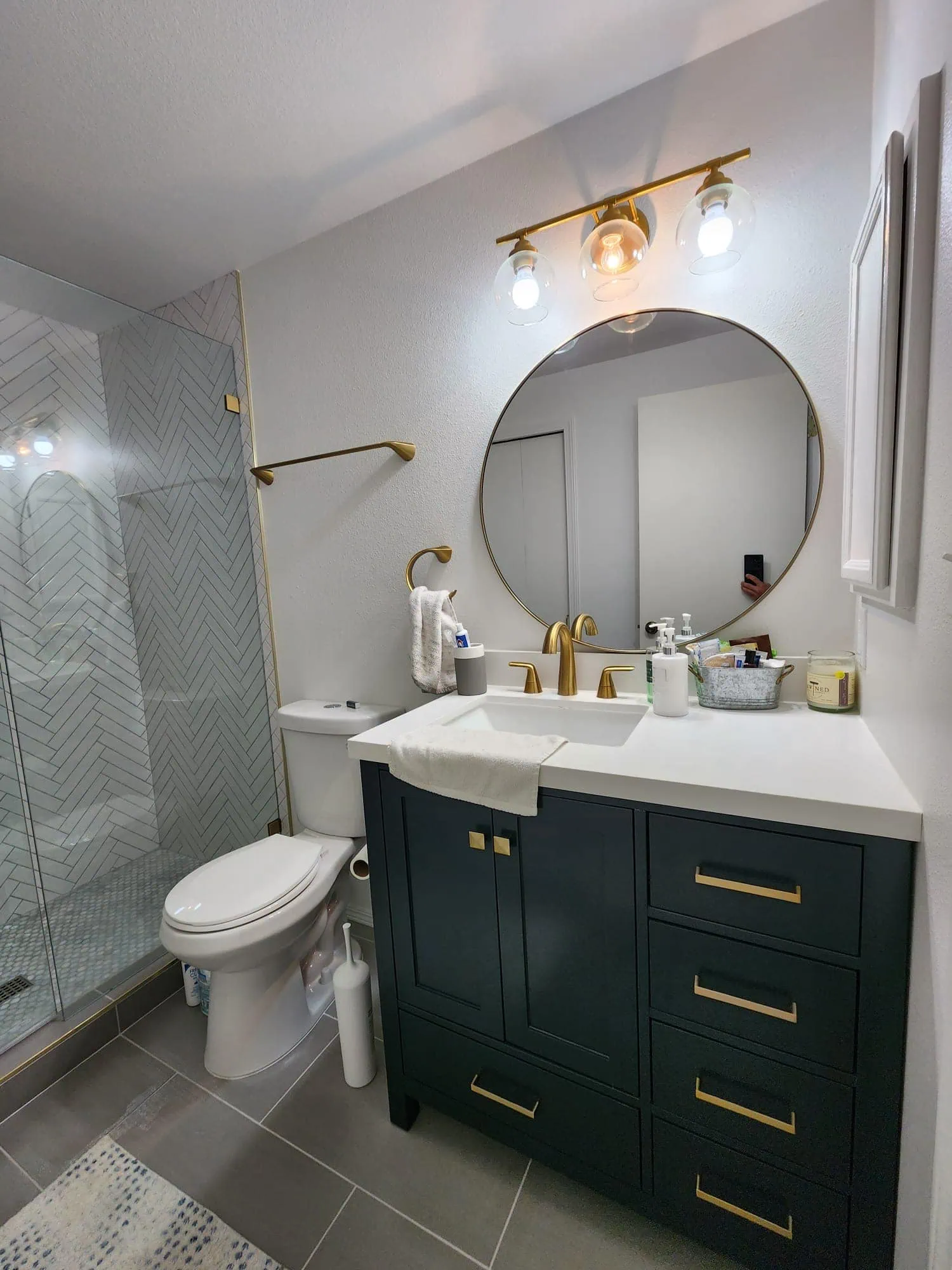 Seattle bathroom remodel featuring a navy vanity with gold hardware, a round mirror, modern lighting, and a herringbone tile shower.