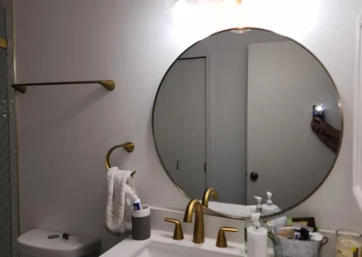 Seattle bathroom remodel featuring a modern vanity with a white quartz countertop, gold fixtures, a round mirror, and elegant lighting.
