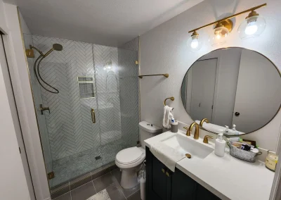 Seattle bathroom remodel featuring a navy vanity with gold hardware, a round mirror, modern lighting, and a glass-enclosed herringbone tile shower.