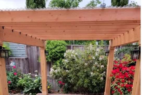 The front of a modern cabin with a gravel driveway, wooden deck, and metal roof, surrounded by forest trees.