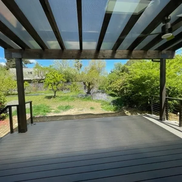 Modern covered patio with a transparent pergola roof, black metal railing, and composite decking, offering a scenic backyard view in Seattle. Designed for style and durability by expert patio installers in Seattle.