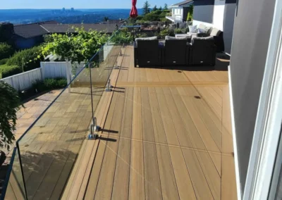 Premium composite deck featuring a diagonal board pattern, frameless glass railings, and a wood-paneled ceiling with integrated lighting.
