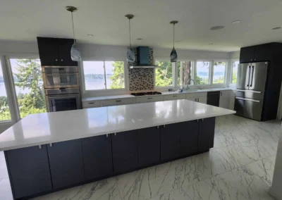 Modern kitchen with a large dark island, white quartz countertops, marble flooring, and waterfront views.