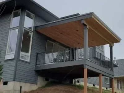 Mercer-Island-home-extension-with-covered-deck-and-railing Modern elevated deck with a covered roof and black metal railing, supported by wooden posts on a sloped yard in Mercer Island. Built by expert deck contractors in Seattle for stability and style.