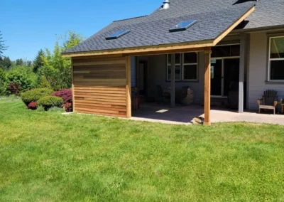 Completed covered patio addition with cedar privacy wall, shingled roof, and skylights integrated into a gray Redmond home, viewed from the lawn