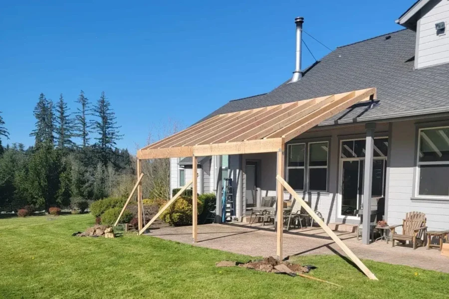redmond-covered-patio-framing-construction-progress Covered patio addition mid-construction with exposed wood framing and temporary supports against a gray Redmond home